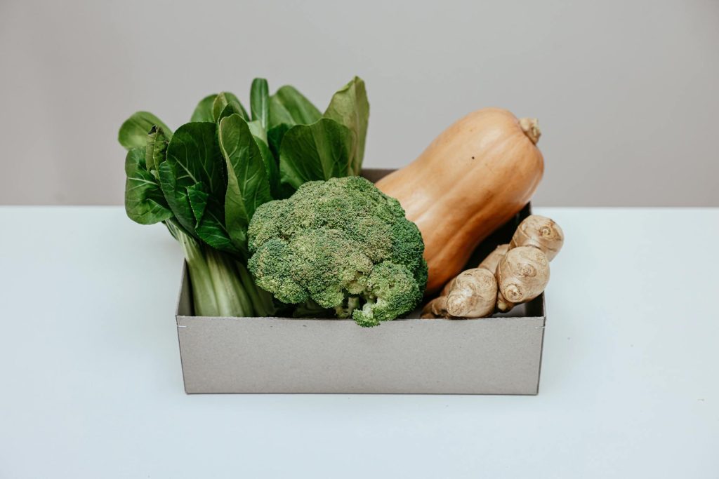 Box of fresh vegetables including broccoli, ginger, and squash on a white table.