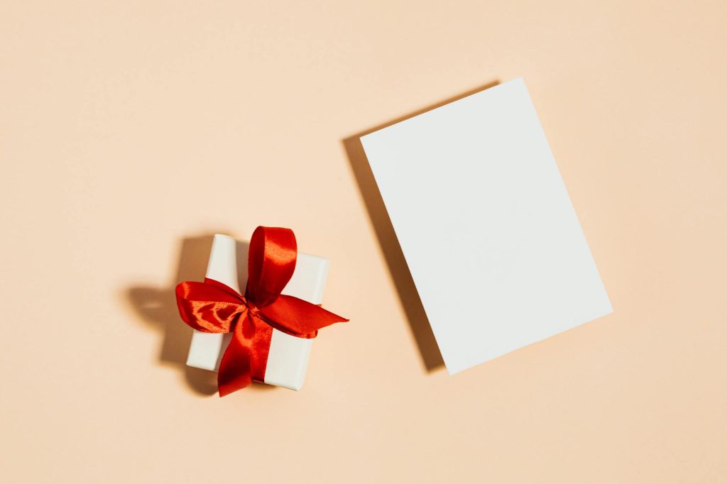 A gift box wrapped with a red ribbon beside a blank postcard on a pastel background.