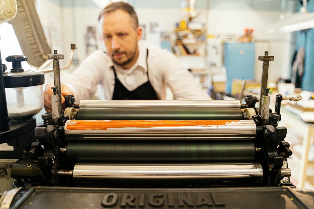 A focused man operates a vintage printing press in a well-lit workshop, highlighting traditional craftsmanship.