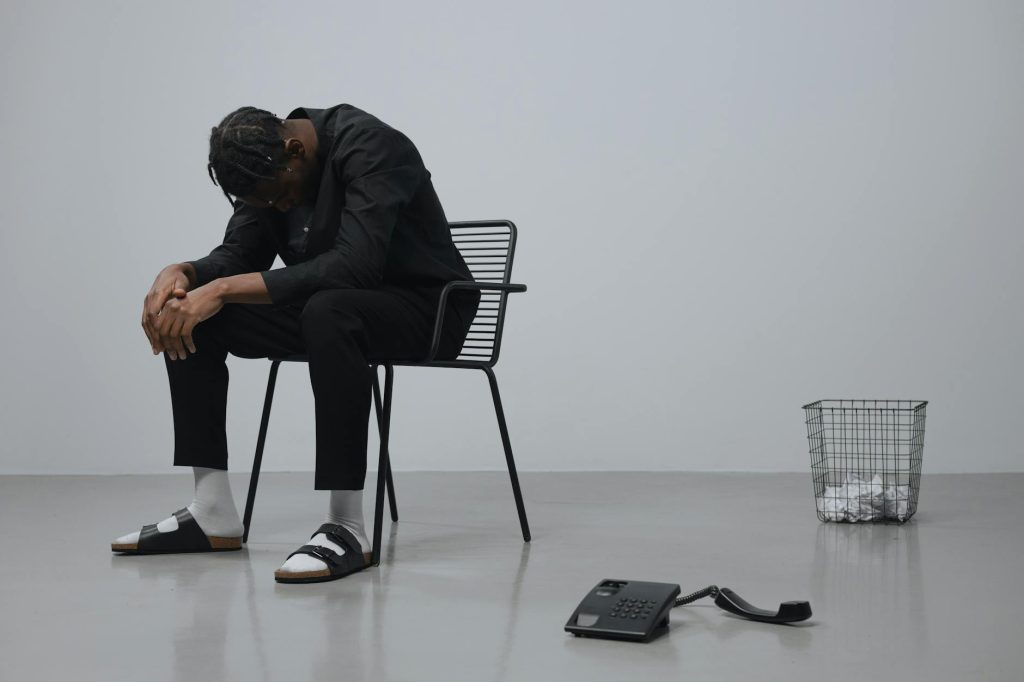 A man in black outfit sitting in a minimalist room, looking tired and overworked.