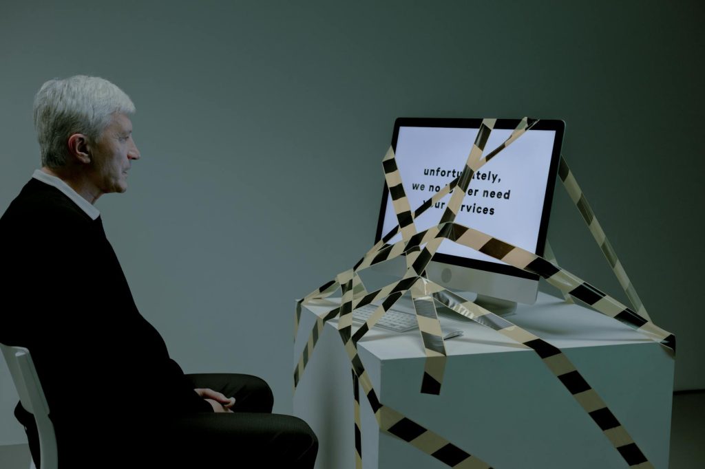 Elderly man confronted with job loss message on computer, symbolizing rejection.