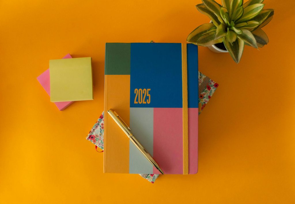 Flat lay of a vibrant 2025 planner with a pen and green plant on yellow background.