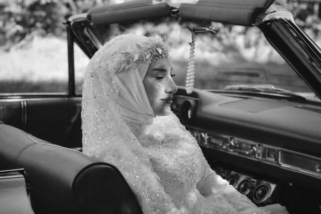 A serene bride in a detailed gown sits in a vintage convertible.