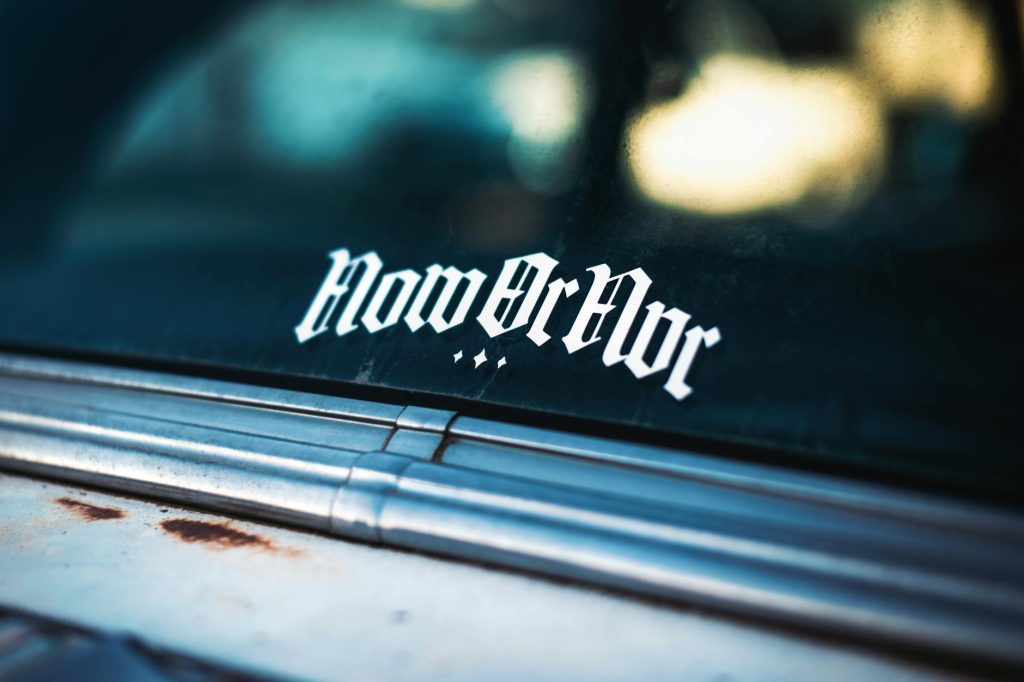 Detailed shot of a vintage car window with a unique 'Now or Nvr' sticker in an urban setting.