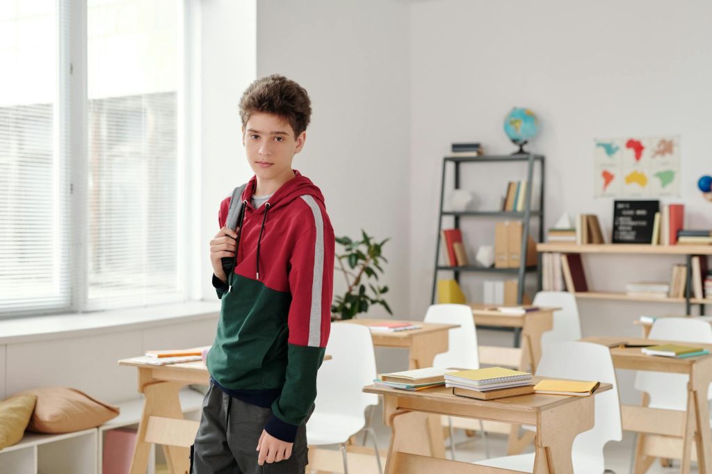 A teenage boy in a hoodie stands in a bright classroom holding a backpack.