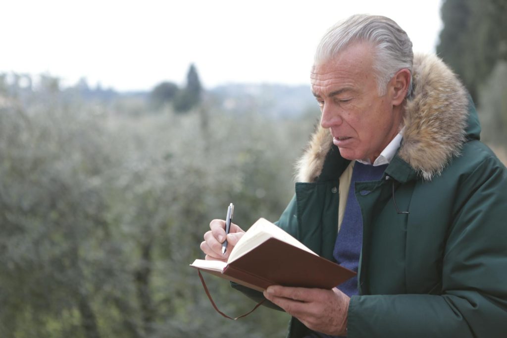Elderly man writing in a notebook while outside in winter clothing.