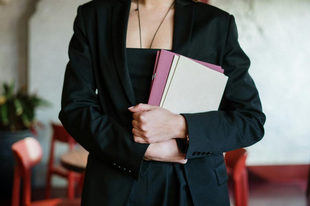 A woman in a black suit holding files, standing in a stylish restaurant interior with red chairs.