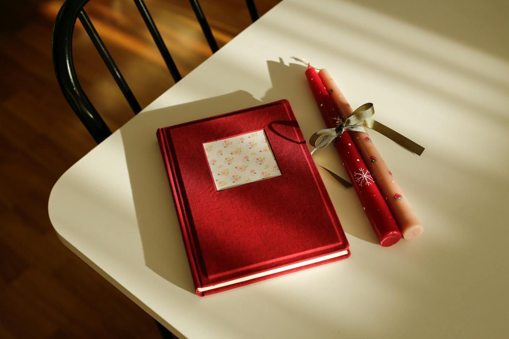 Red journal and decorative candles on table lit by warm sunlight, creating a cozy, inviting atmosphere.