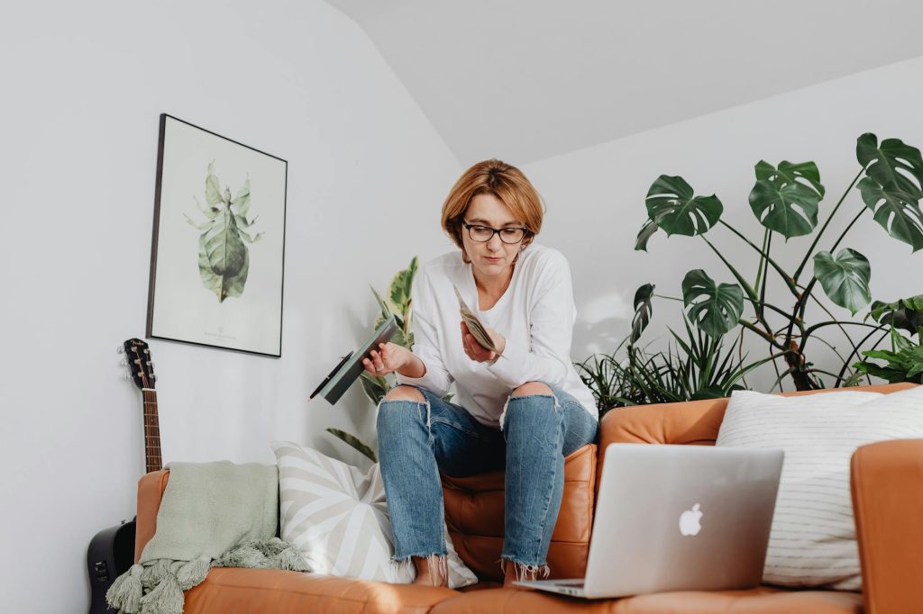 Woman on a cozy sofa using her laptop and smartphone for a video call amidst indoor plants.