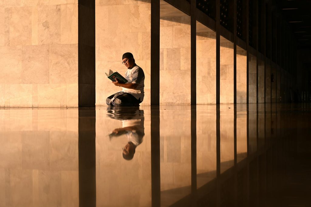 A man sits reading a book in a reflective corridor of a Jakarta mosque during golden hour.