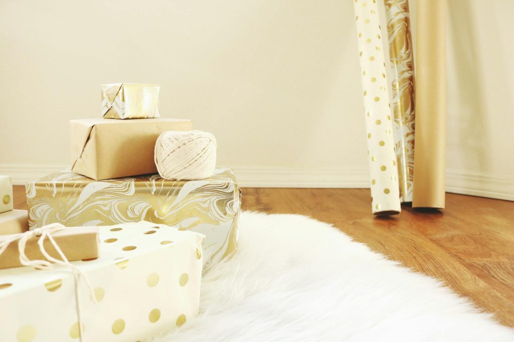 Stack of beautifully wrapped gifts in gold and white tones on wooden floor.