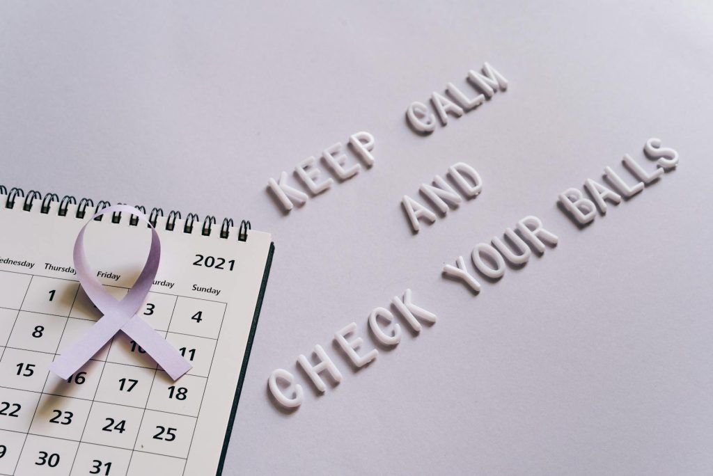 Flat lay image of a calendar with a purple ribbon and the text 'Keep Calm and Check Your Balls' for testicular cancer awareness.