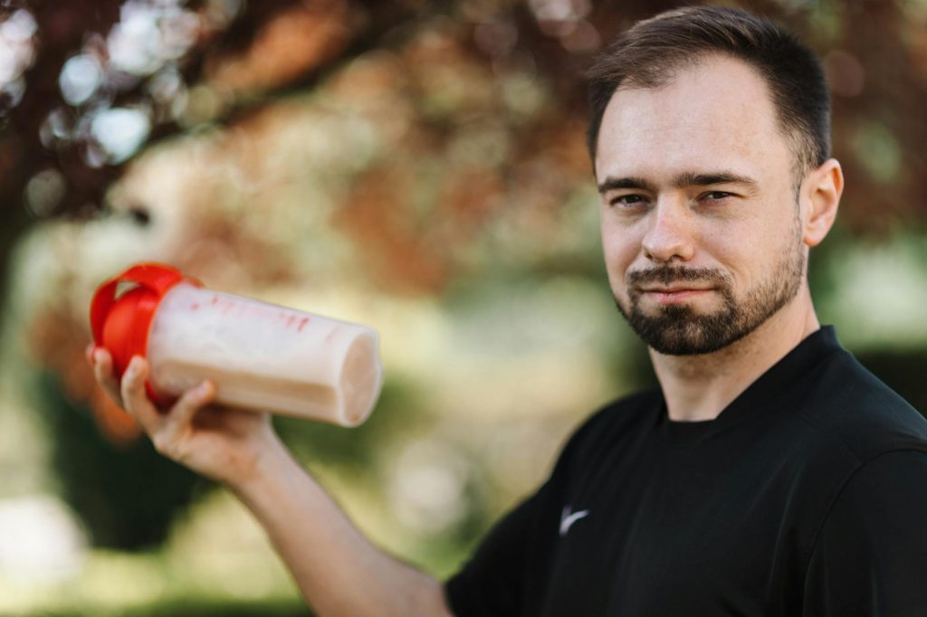 Adult man holding a protein shake outdoors during the day.