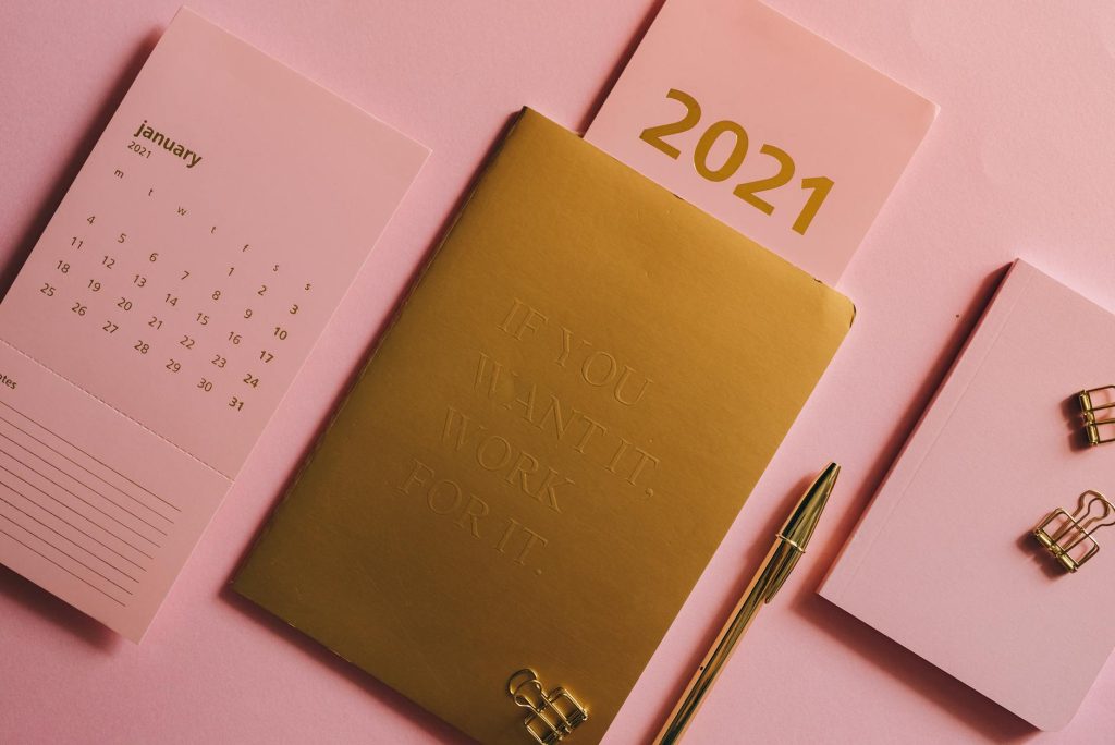 A minimalist desktop with a 2021 calendar, golden notebook, and stationary on a pink background.