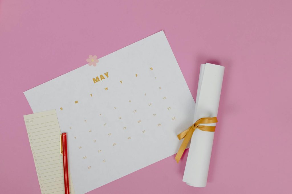 Flat lay of a May calendar with a pen and paper roll on pink background.