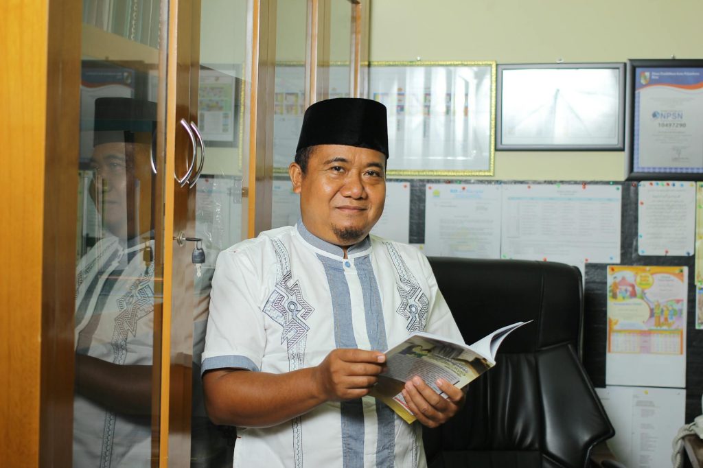 A confident Indonesian teacher holding a book in his office, showcasing education and professionalism.