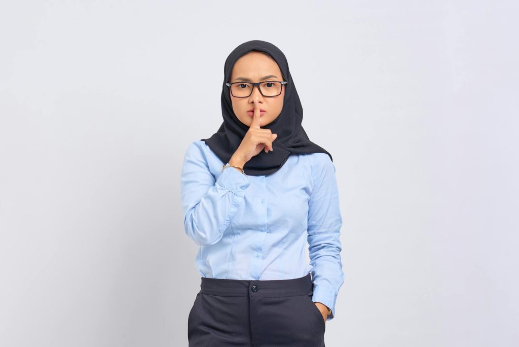 Asian Muslim woman in hijab making a quiet gesture with a serious expression, wearing eyeglasses against a white background.