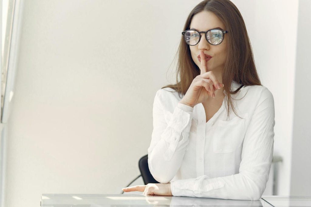 Young woman with long hair in trendy clothes sitting at table and doing shh gesture asking to stay quiet in contemporary office