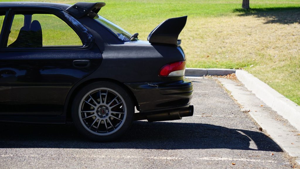 Black Subaru Impreza with spoiler parked outdoors, sunlight casting shadows.