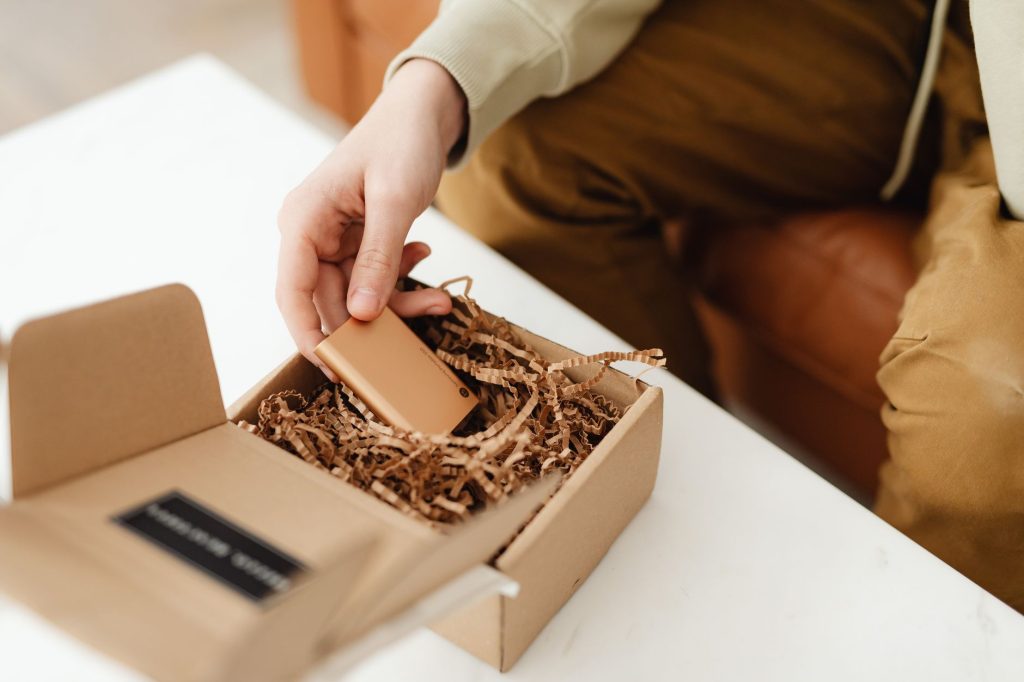 A person unboxes a small item from a cardboard package filled with crinkle paper, indoors.